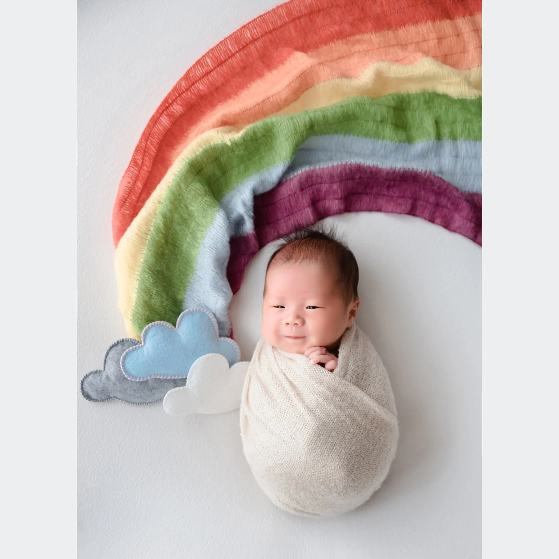 Sleeping newborn in a cream swaddle beneath a rainbow made from colourful fabric beside felt clouds on a white backdrop