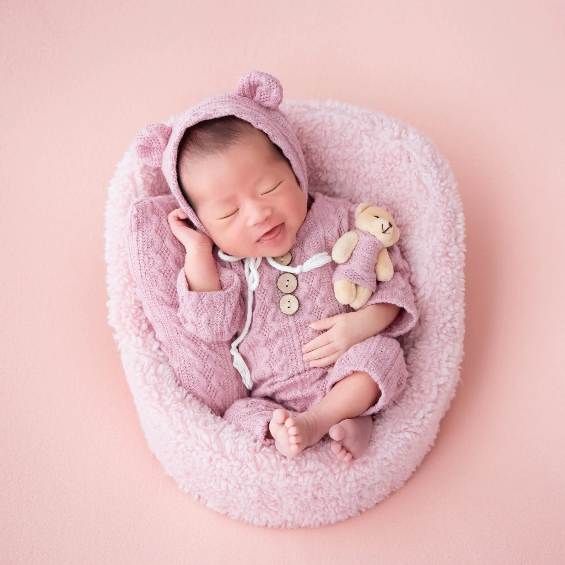 Sleeping newborn in a pink knitted bear hat and romper with a small teddy bear in a pink basket