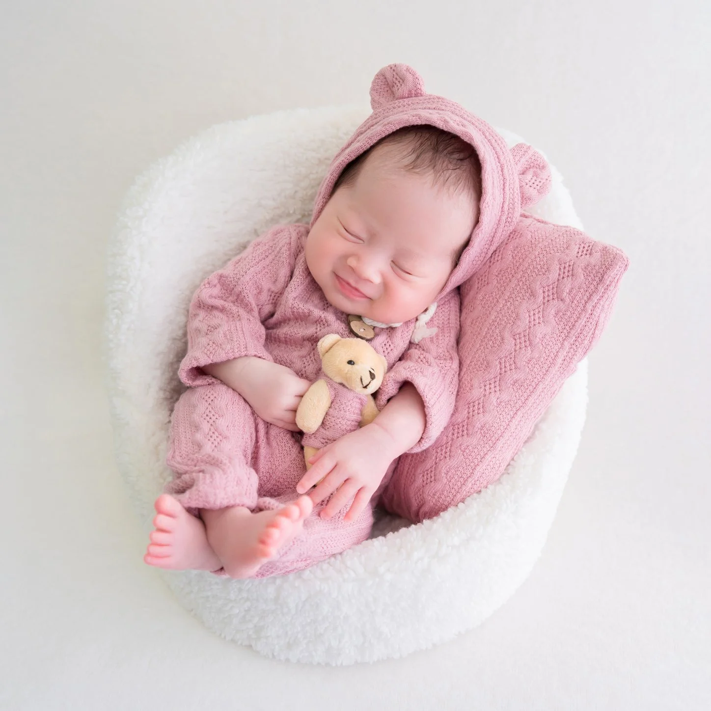 Sleeping newborn in a pink bear outfit holding a tiny teddy bear, curled in a white basket with a pink cushion