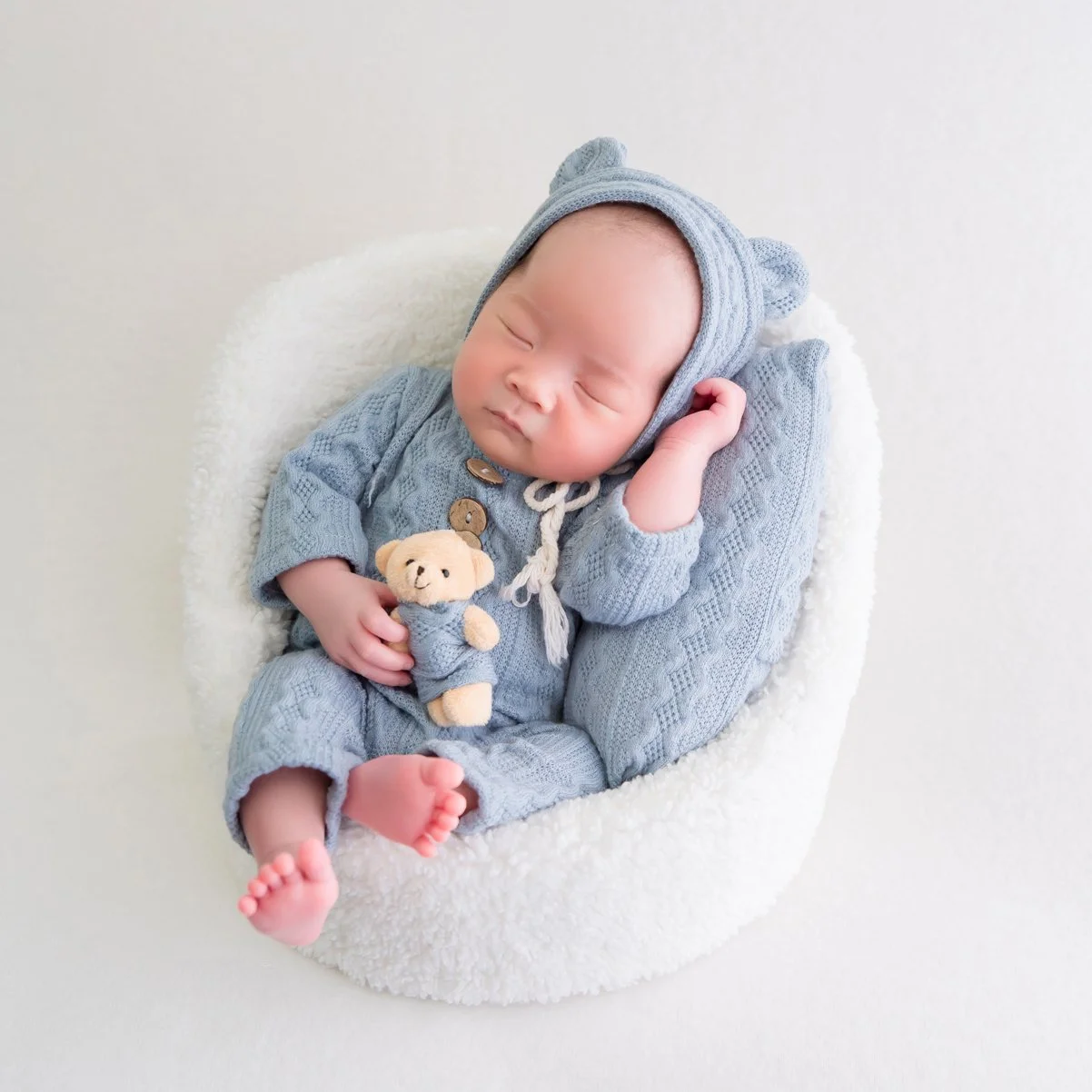 Sleeping newborn in a blue knitted bear hat and romper holding a tiny teddy bear in a white basket