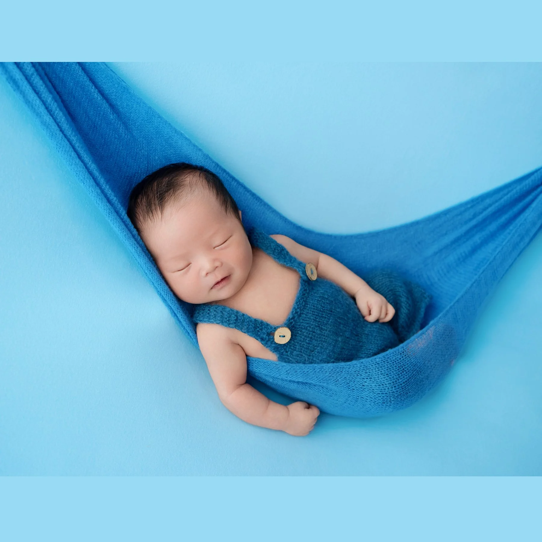 Sleeping newborn in blue knitted overalls reclining in a blue fabric hammock on a matching blue backdrop