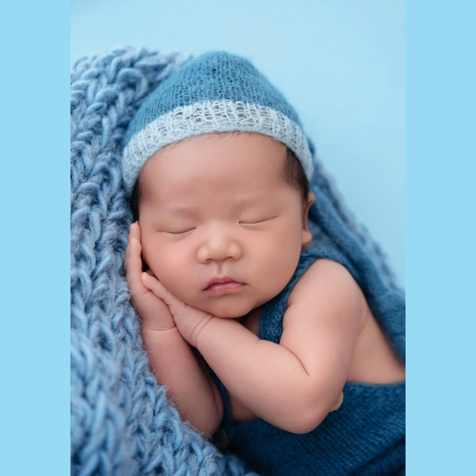 Close-up of a sleeping newborn in a blue knitted hat resting the cheek on both hands against a blue knitted blanket