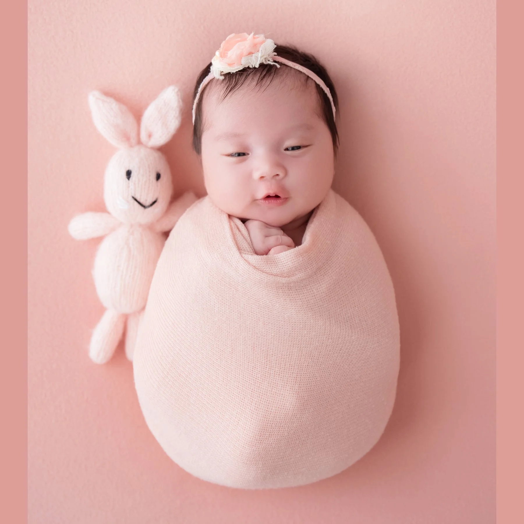 Yawning newborn in a pale pink swaddle and floral headband reclining beside a pink rabbit plush on a pink backdrop