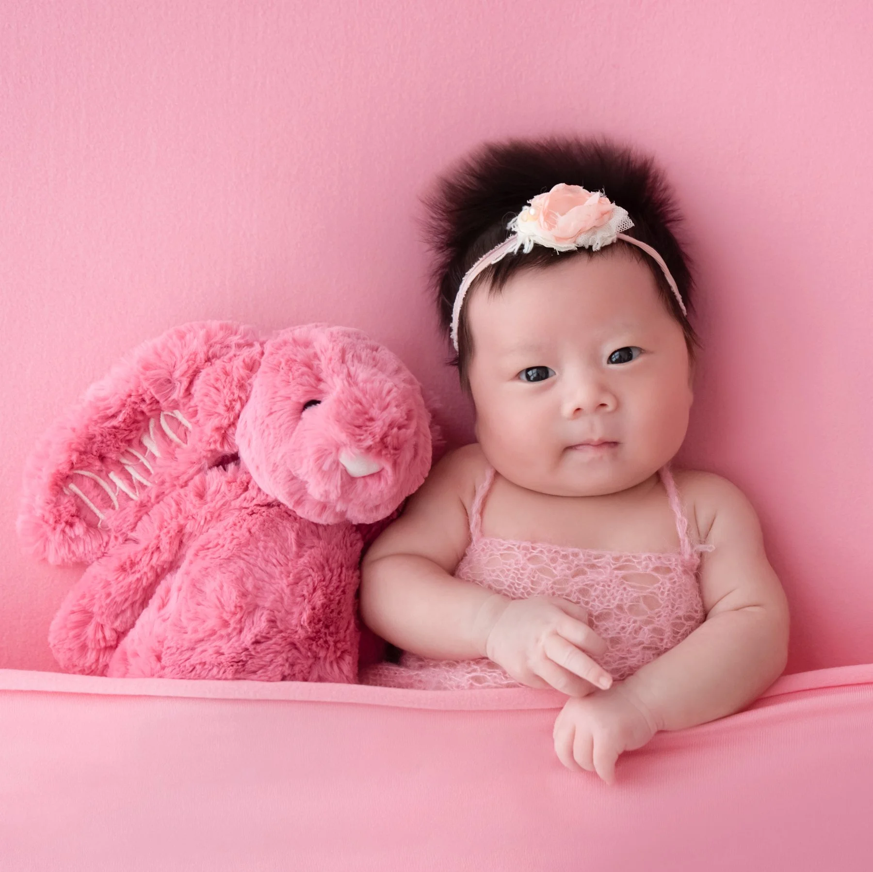 Awake newborn in a pink knitted outfit and floral headband resting on folded arms beside a pink elephant plush on a pink backdrop