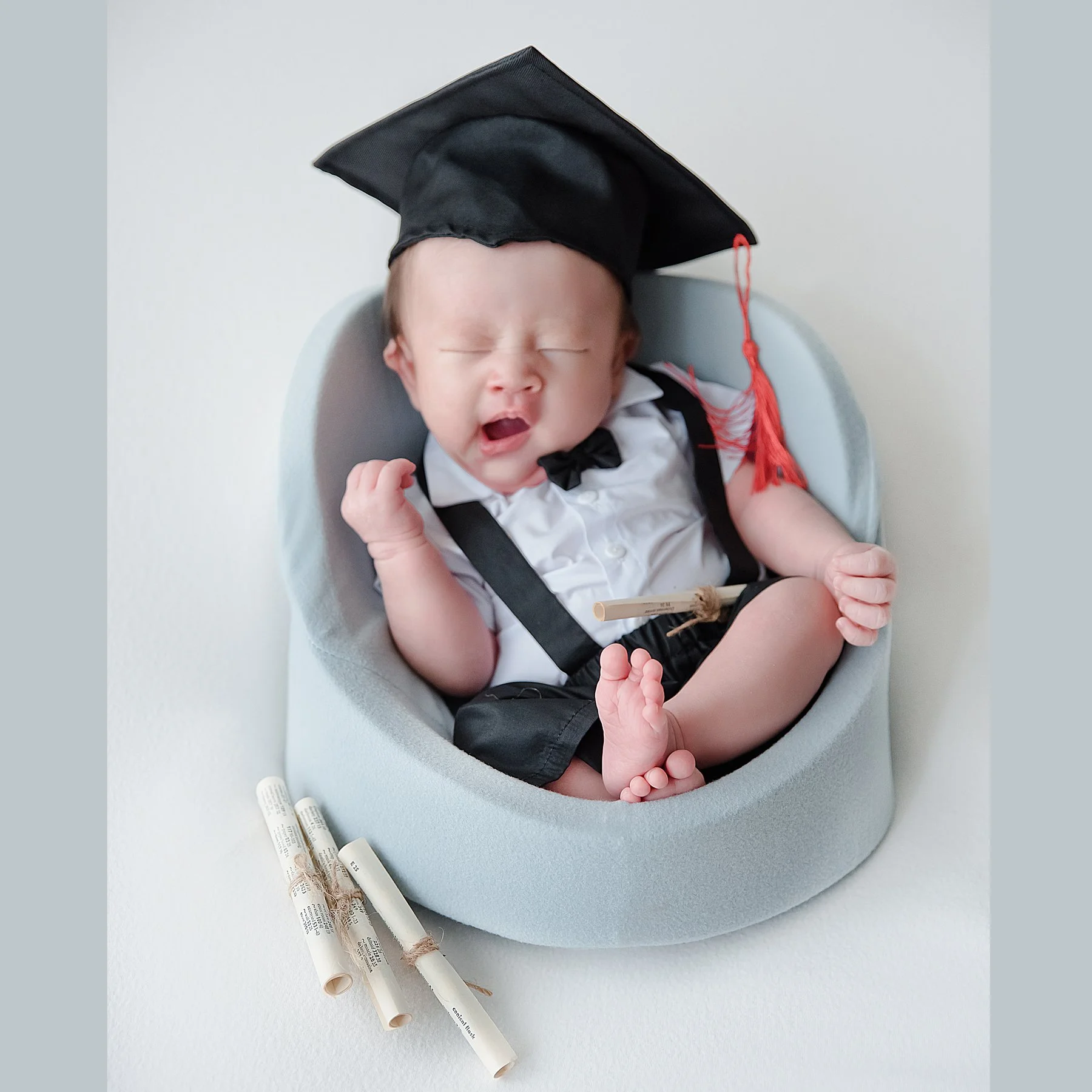 Yawning newborn wearing a graduation cap, white shirt and black bow tie seated in a pale blue basket beside rolled certificates