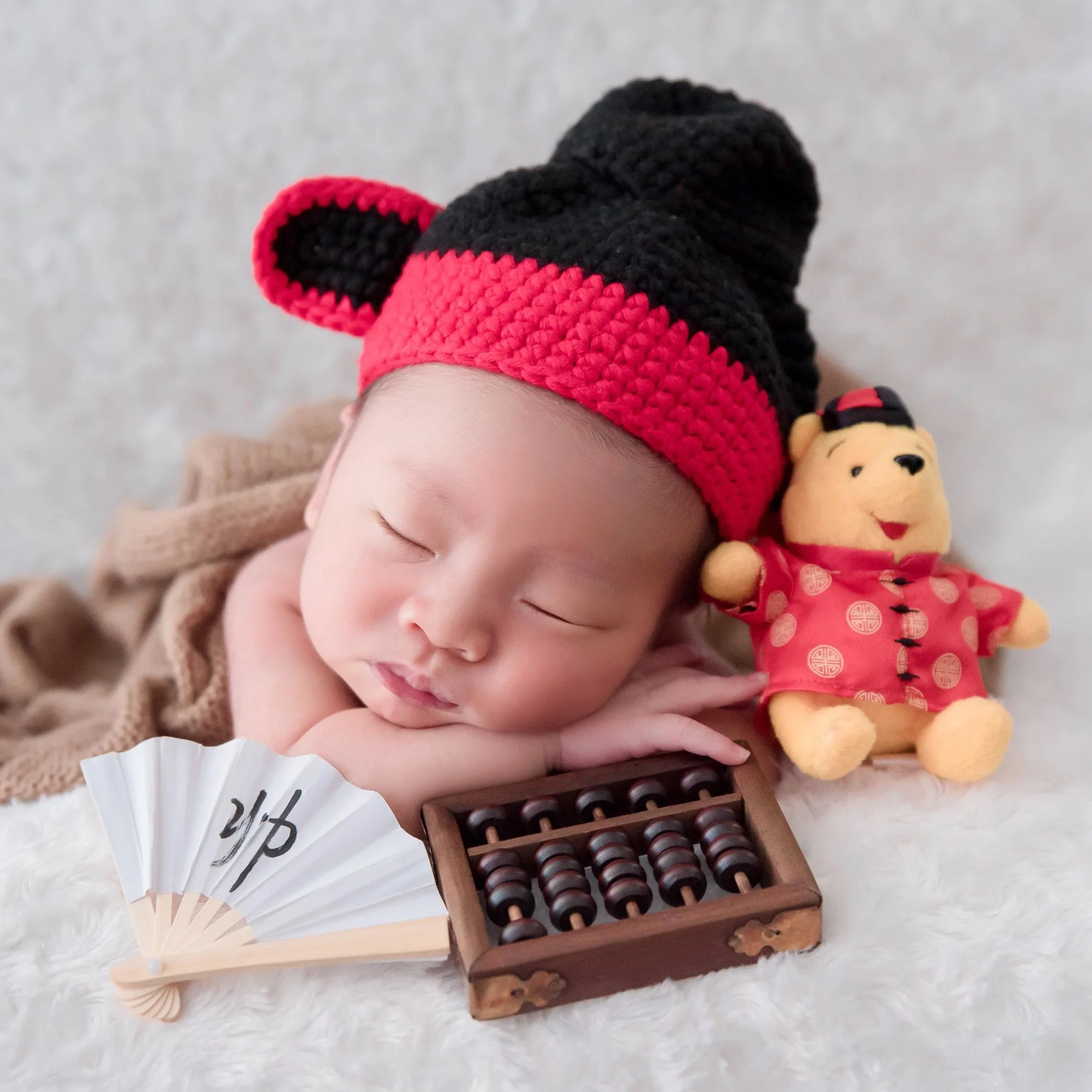 Close-up of a sleeping newborn in a black-and-red Chinese scholar hat resting on folded arms beside a miniature abacus and festive teddy bear