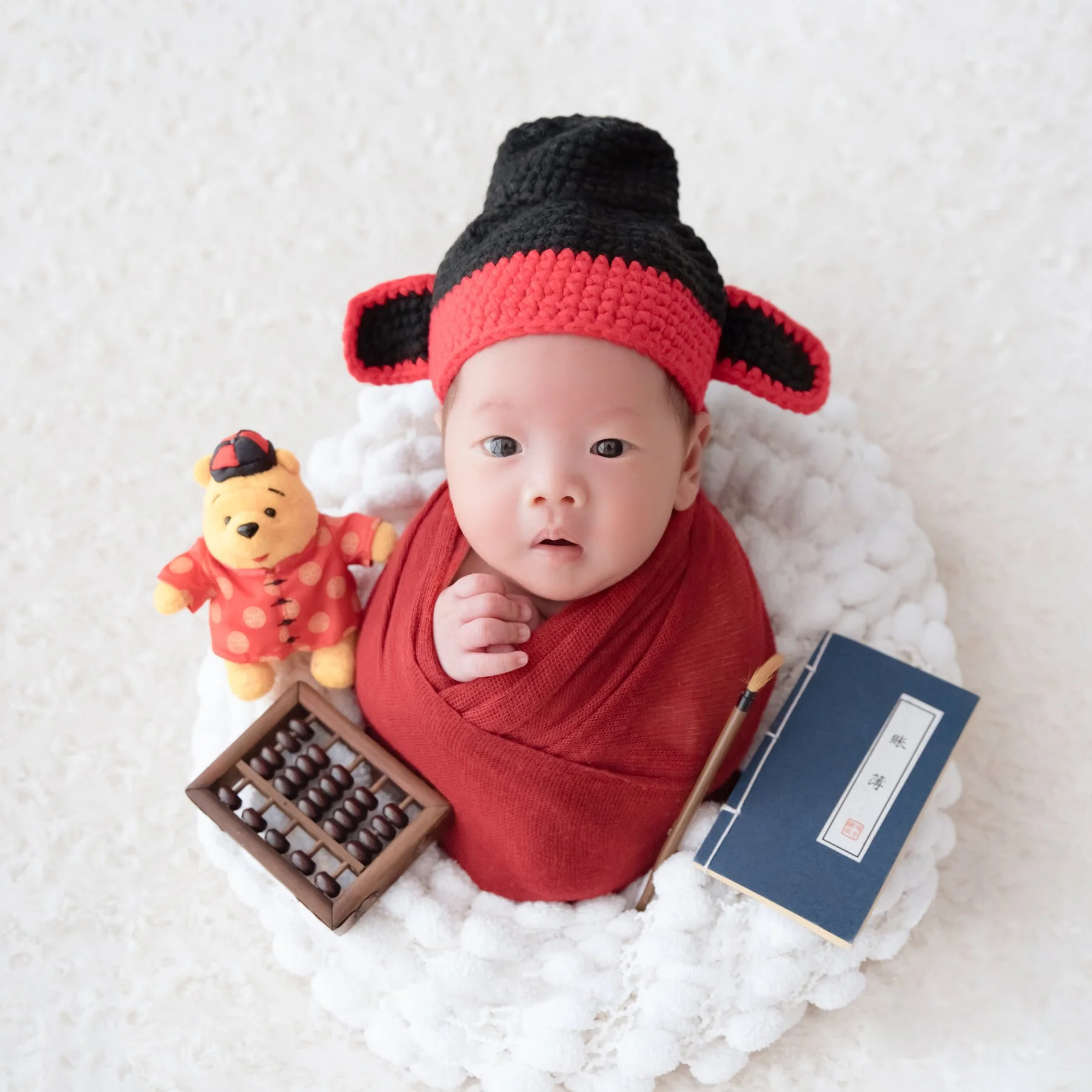 Awake newborn in a red Chinese scholar outfit and black-and-red hat lying on white fur beside a teddy bear, miniature abacus and books