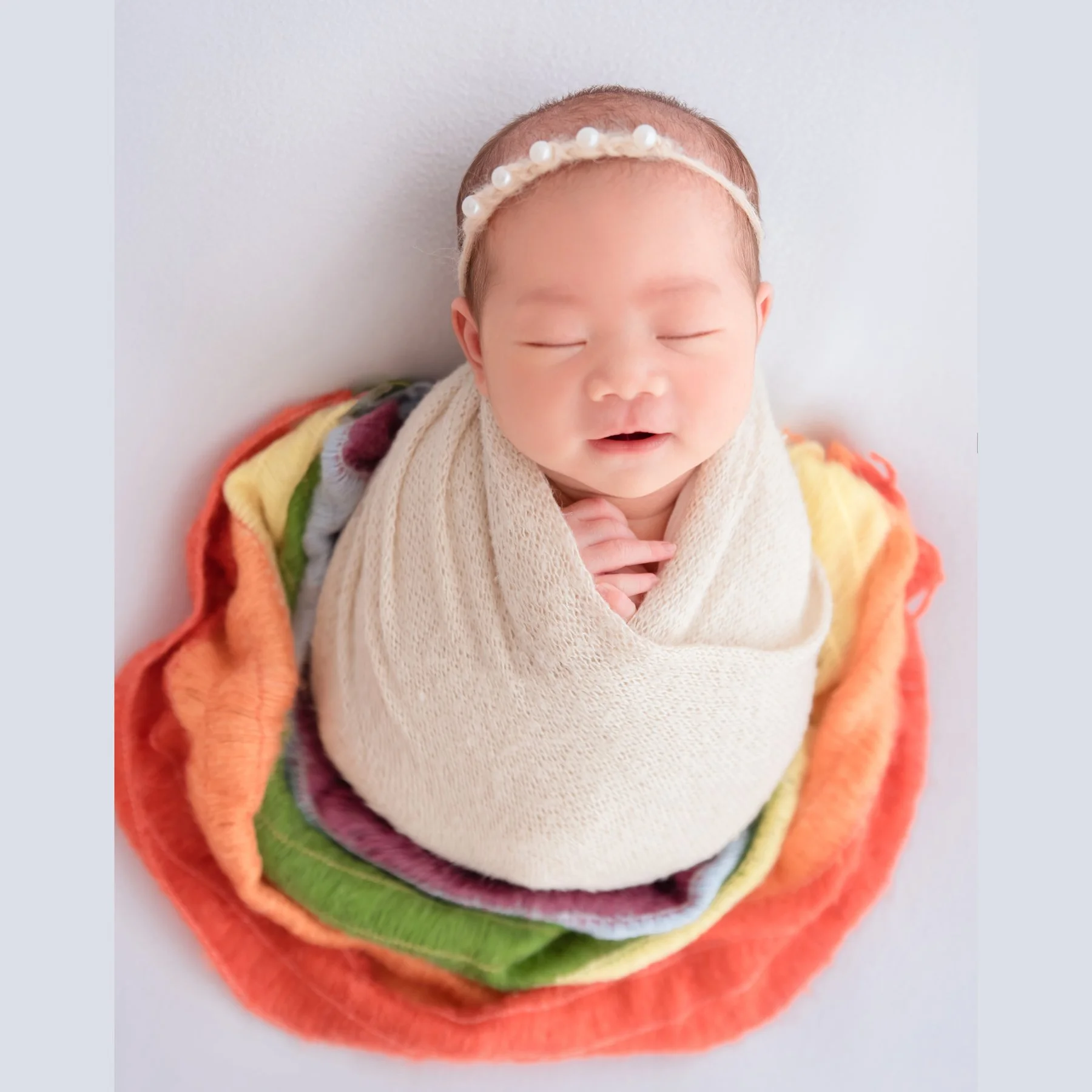 Yawning newborn in a cream swaddle and pearl headband nestled in a circular rainbow of colourful fabric on a white backdrop