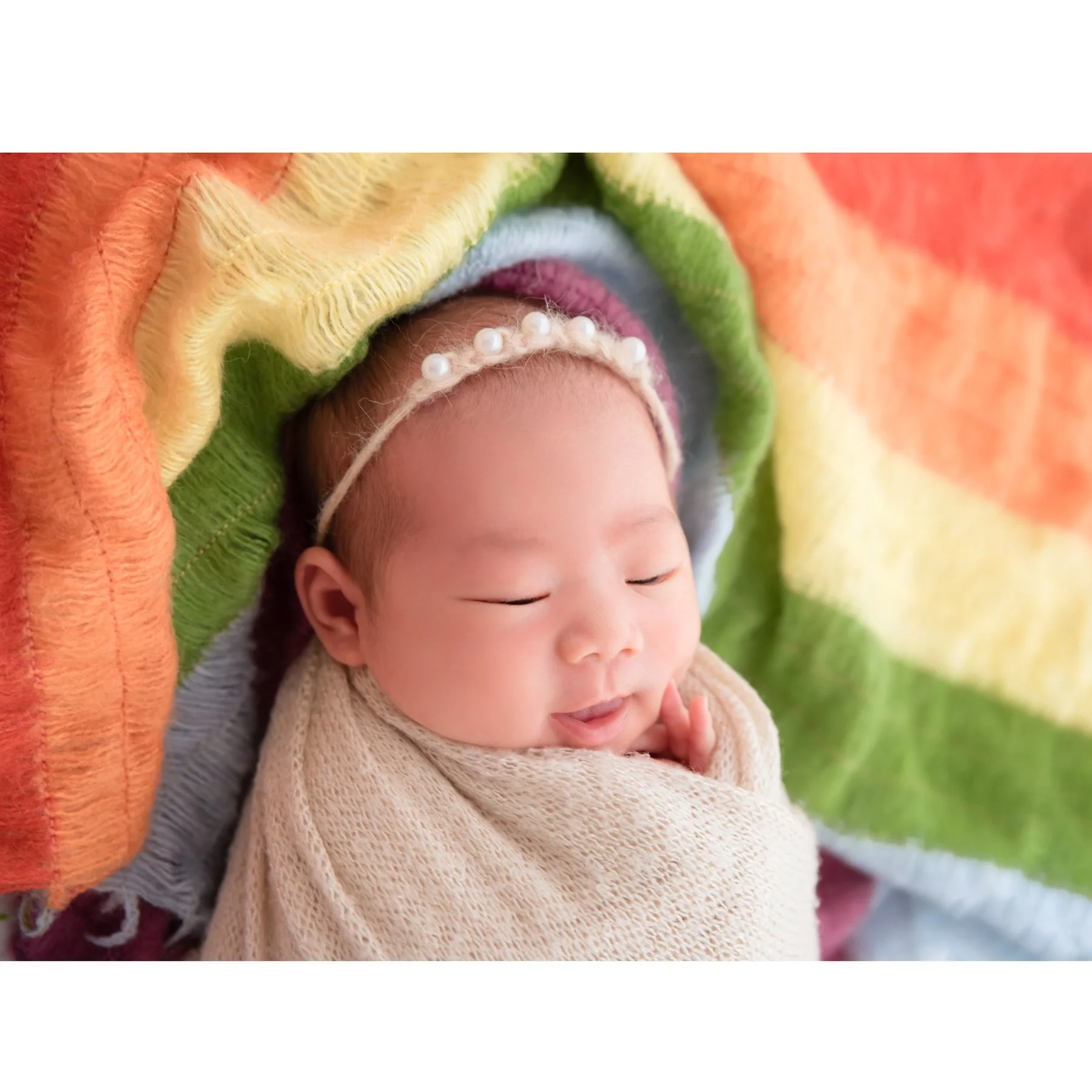 Close-up of a sleeping newborn in a cream swaddle and pearl headband with one hand beside the cheek against colourful rainbow fabric