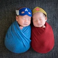 Newborn twins wrapped in blue and red with Captain America and Wonder Woman hats