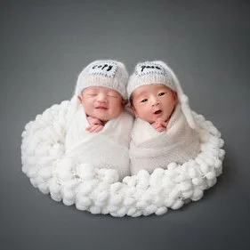 Newborn twins wrapped in white with cloud-patterned knitted hats in a fluffy white basket