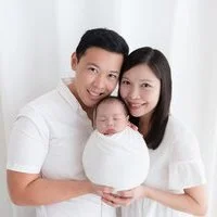 Smiling parents in white holding their sleeping newborn wrapped in white