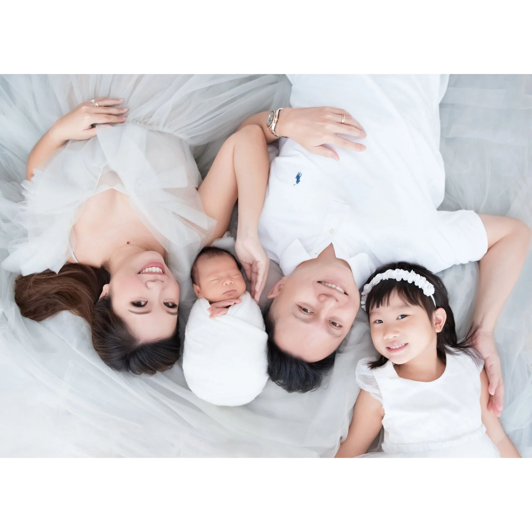 Family of four lying together on a white blanket with their newborn, photographed from above