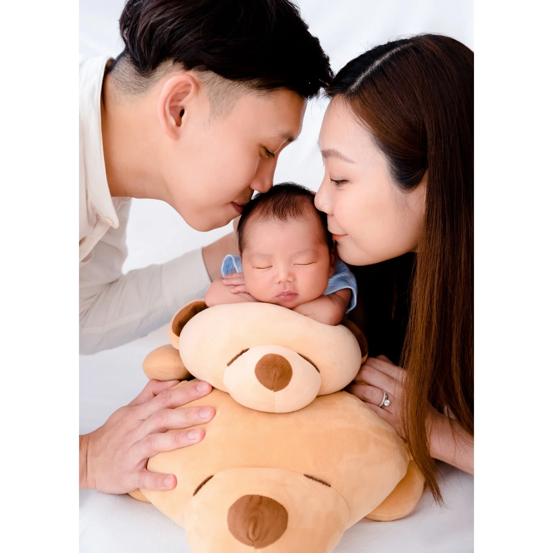Parents kissing their newborn’s head while holding a teddy bear beneath the baby