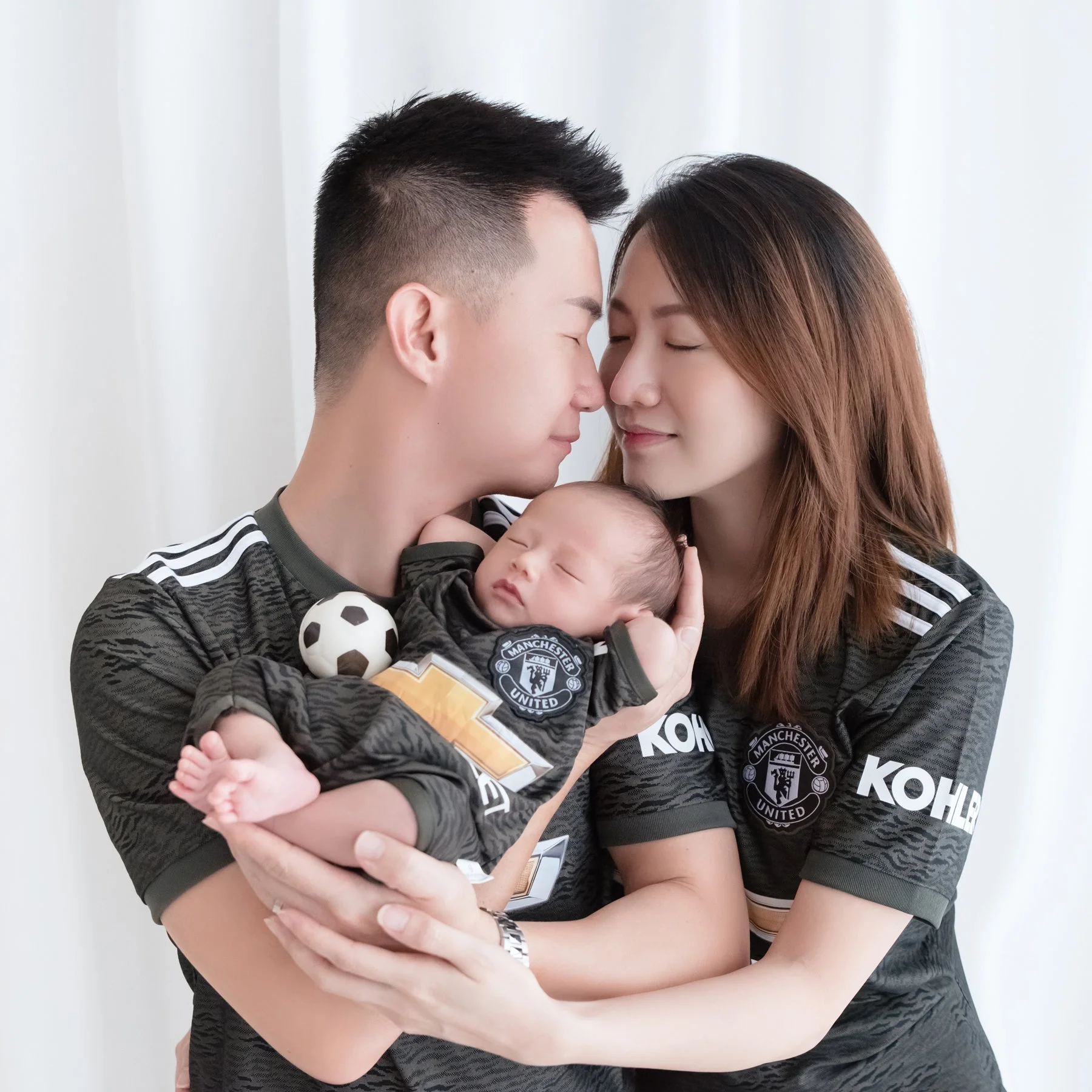 Parents wearing matching football shirts and kissing while holding their newborn