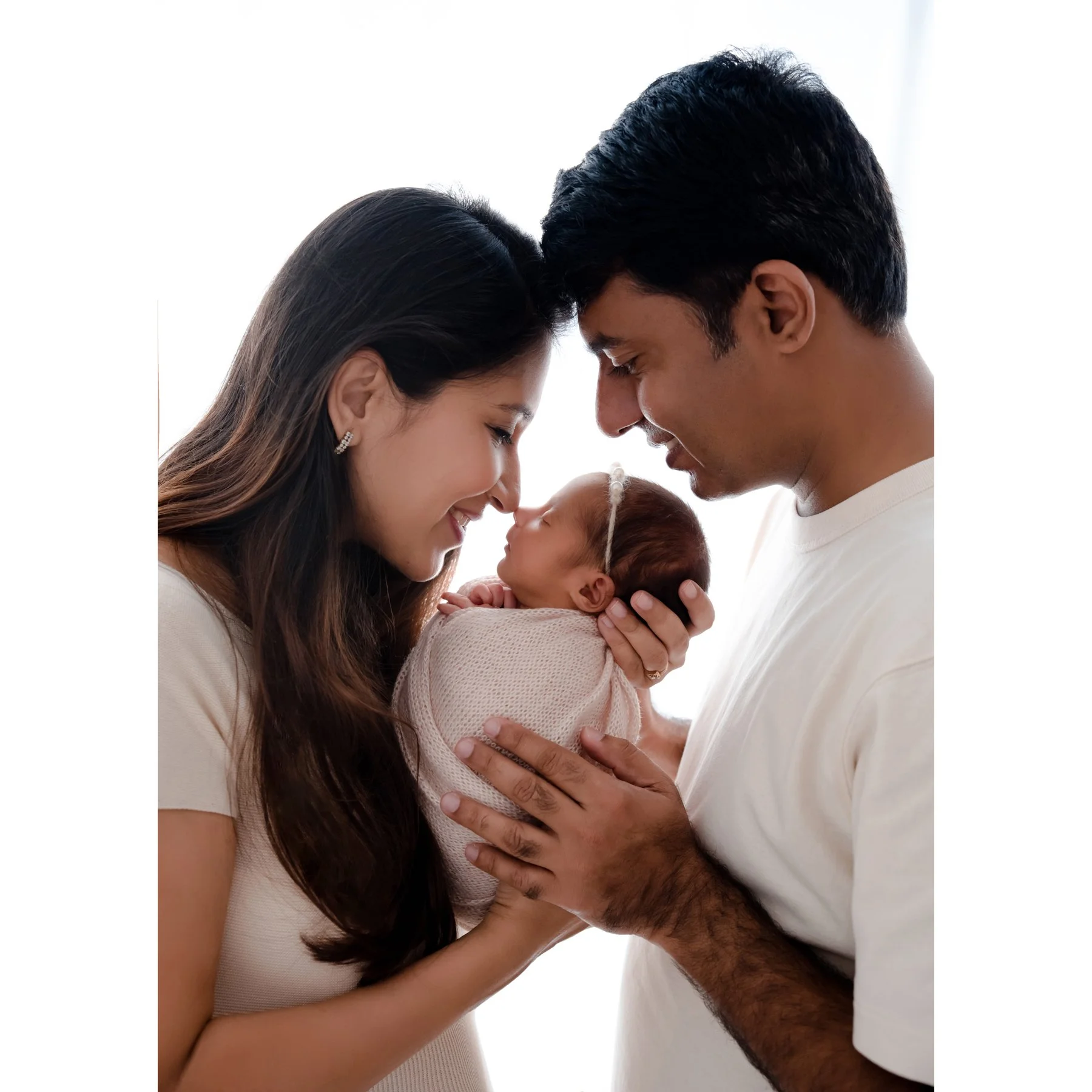 Parents touching foreheads while cradling their newborn wrapped in white