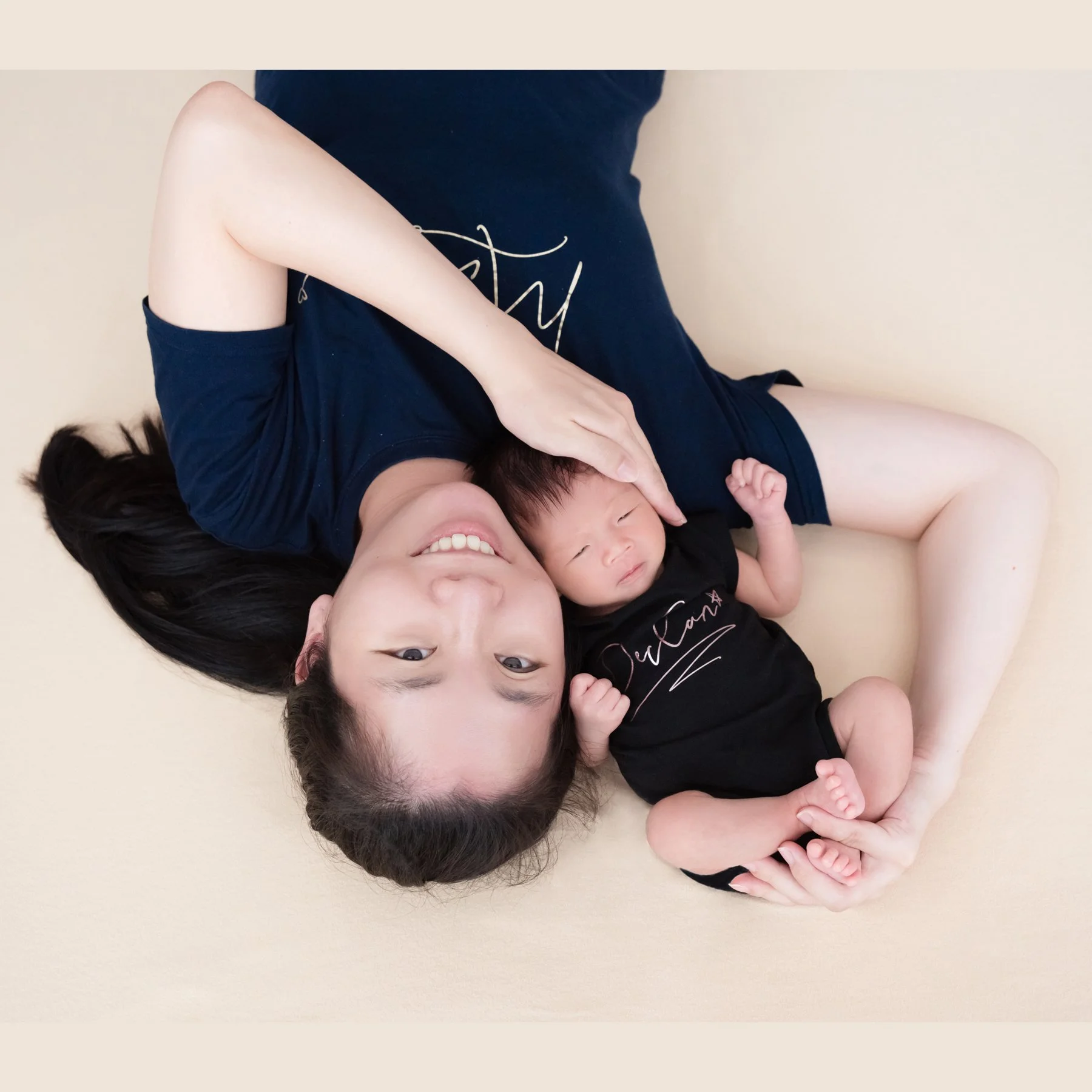Mother in a black dress lying on a cream blanket beside her sleeping newborn
