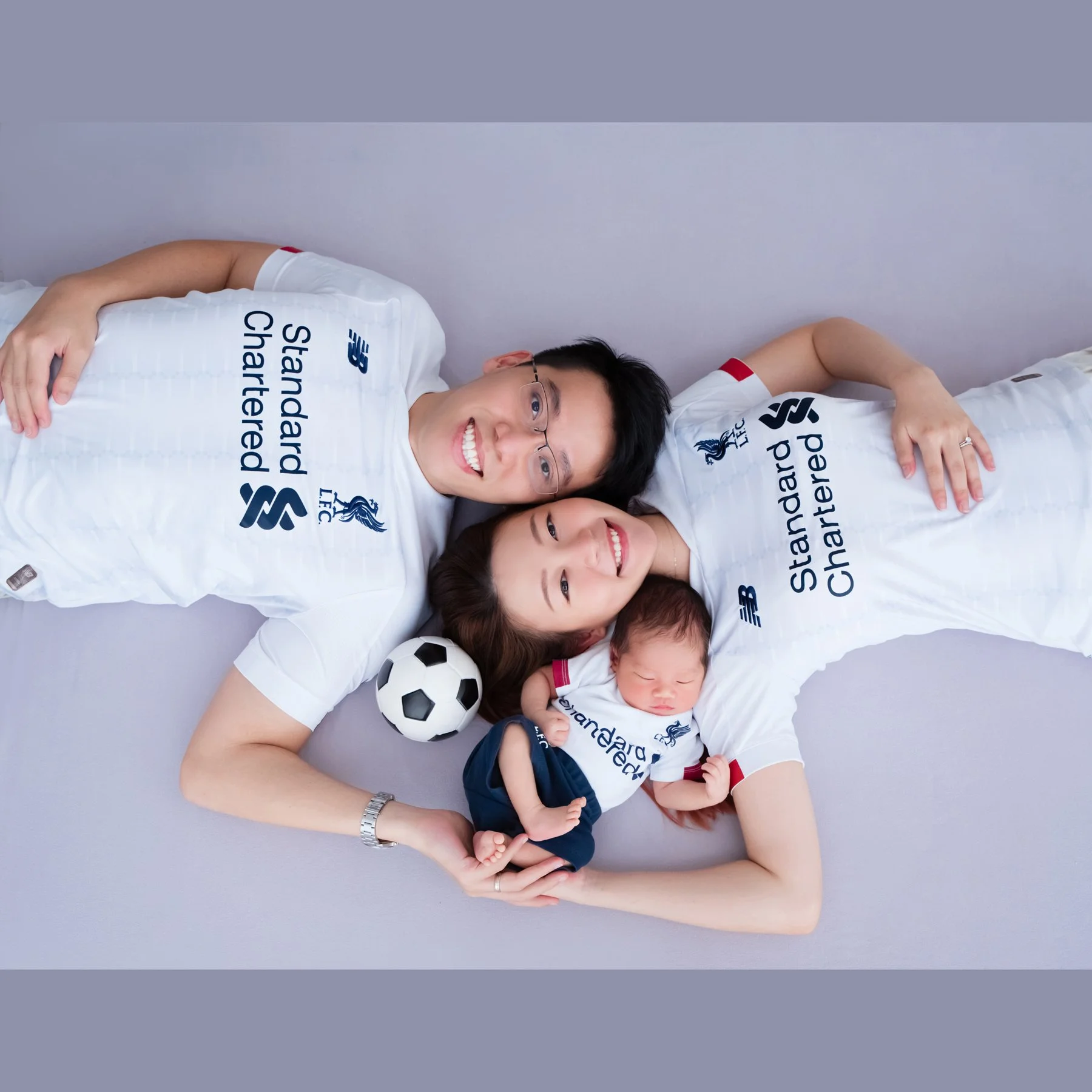 Family lying on a grey blanket in matching white football shirts with their newborn and a football