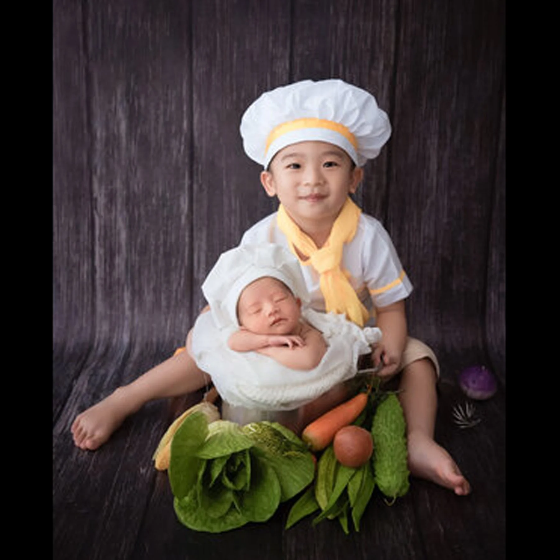 Older brother wearing a chef hat and holding his newborn sibling beside fresh vegetables
