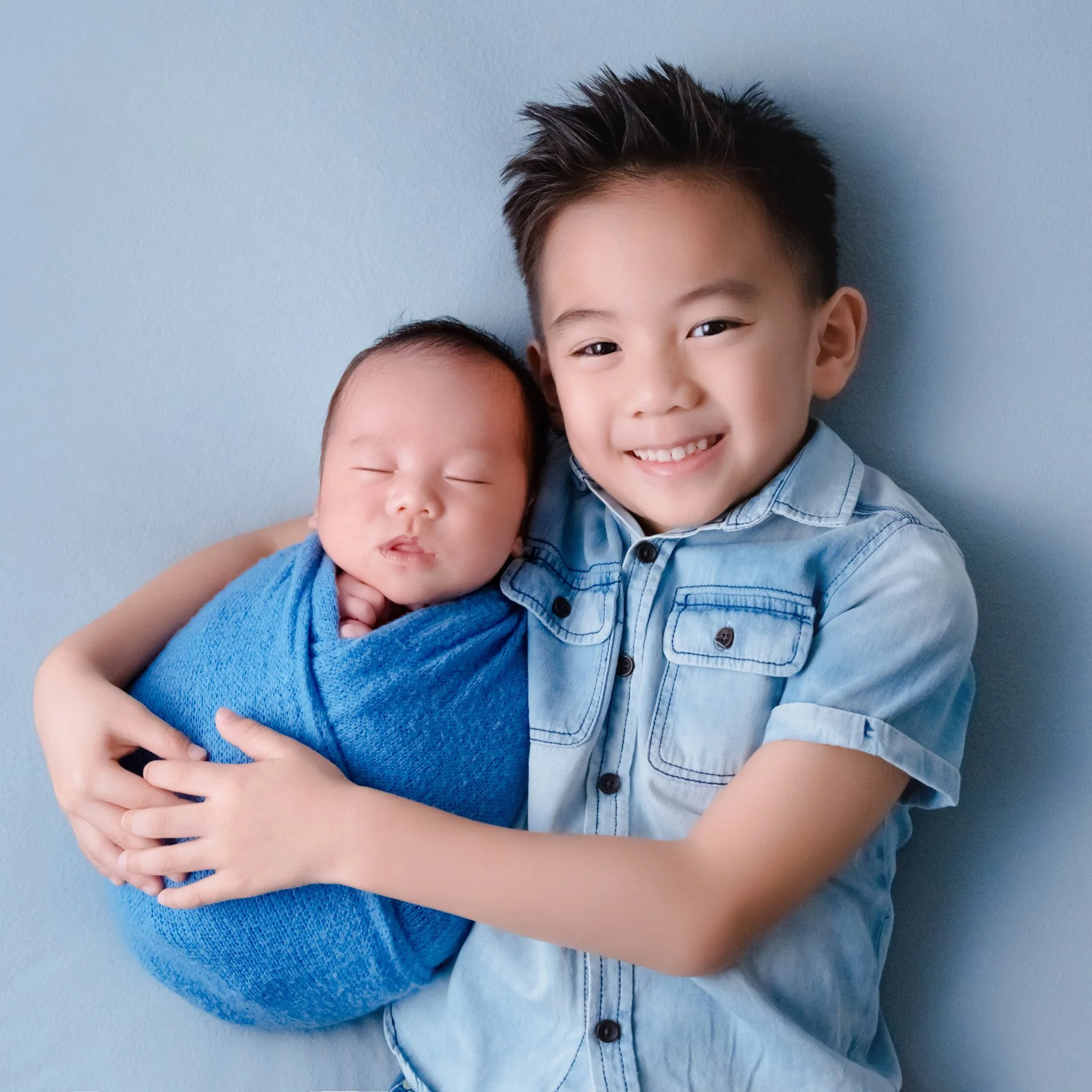 Smiling older brother in a denim shirt holding his newborn sibling wrapped in blue