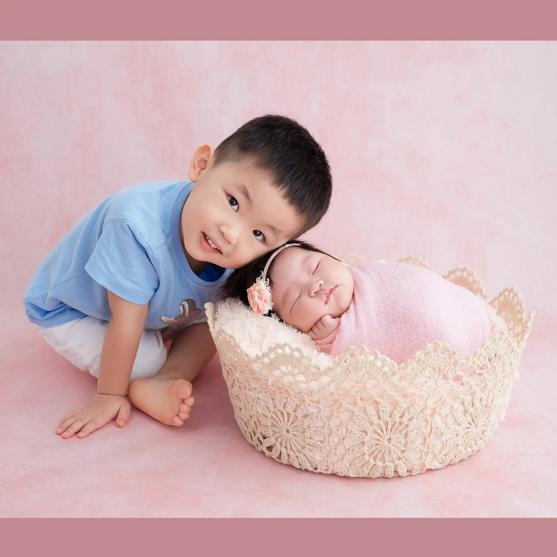Older brother leaning over his newborn sibling in a cream basket against a pink backdrop