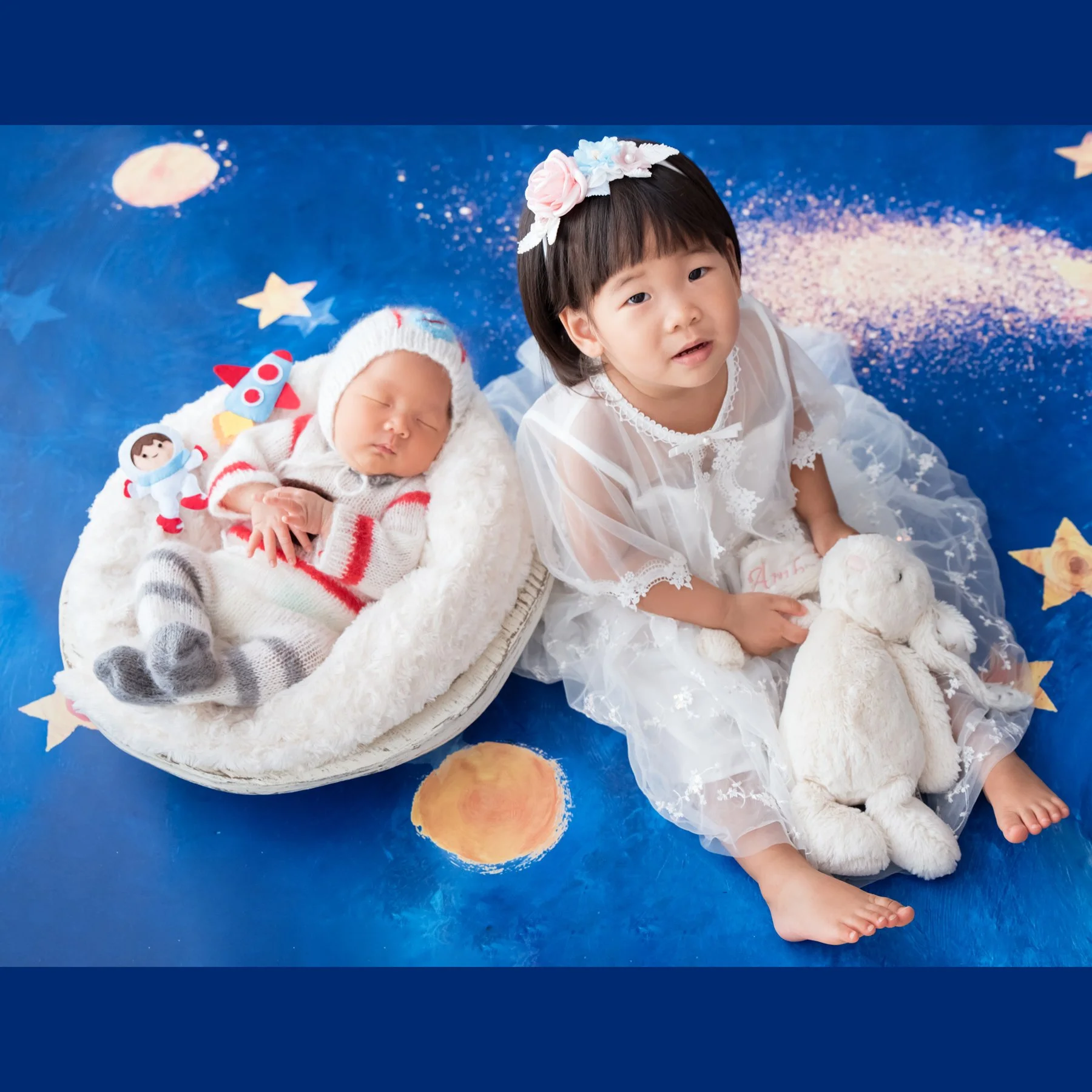 Older sister sitting beside her newborn sibling in an astronaut-themed scene with planets and stars