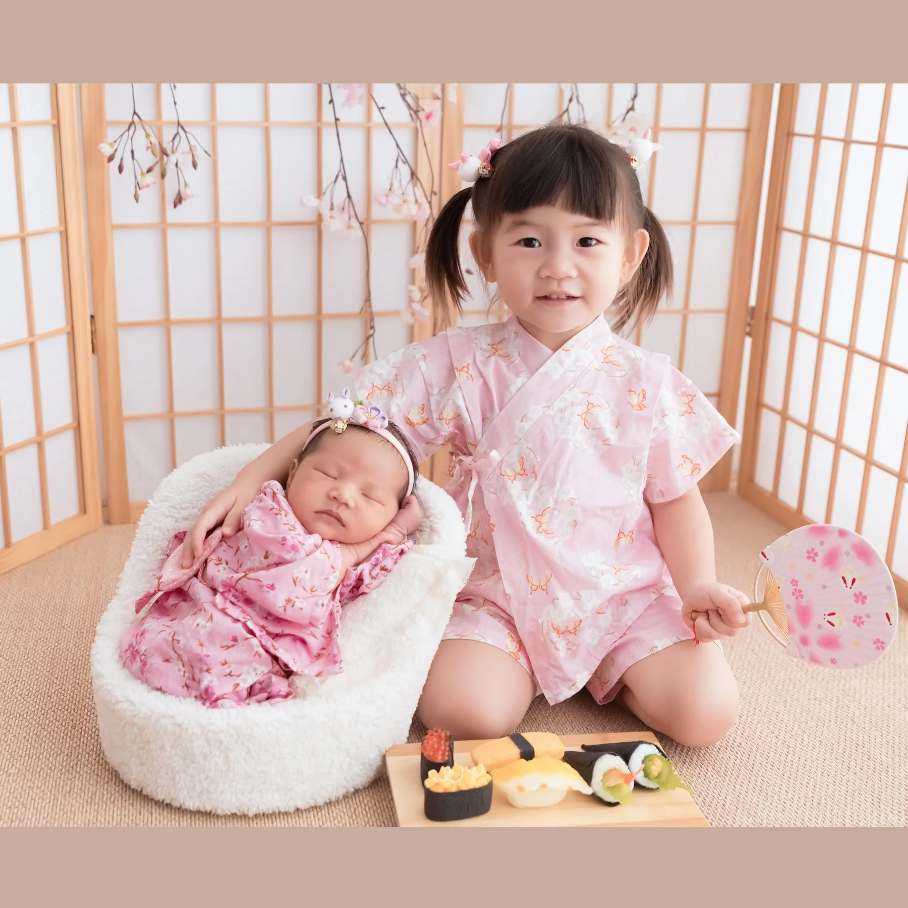 Older sister in a pink kimono sitting beside her newborn sibling in a white basket