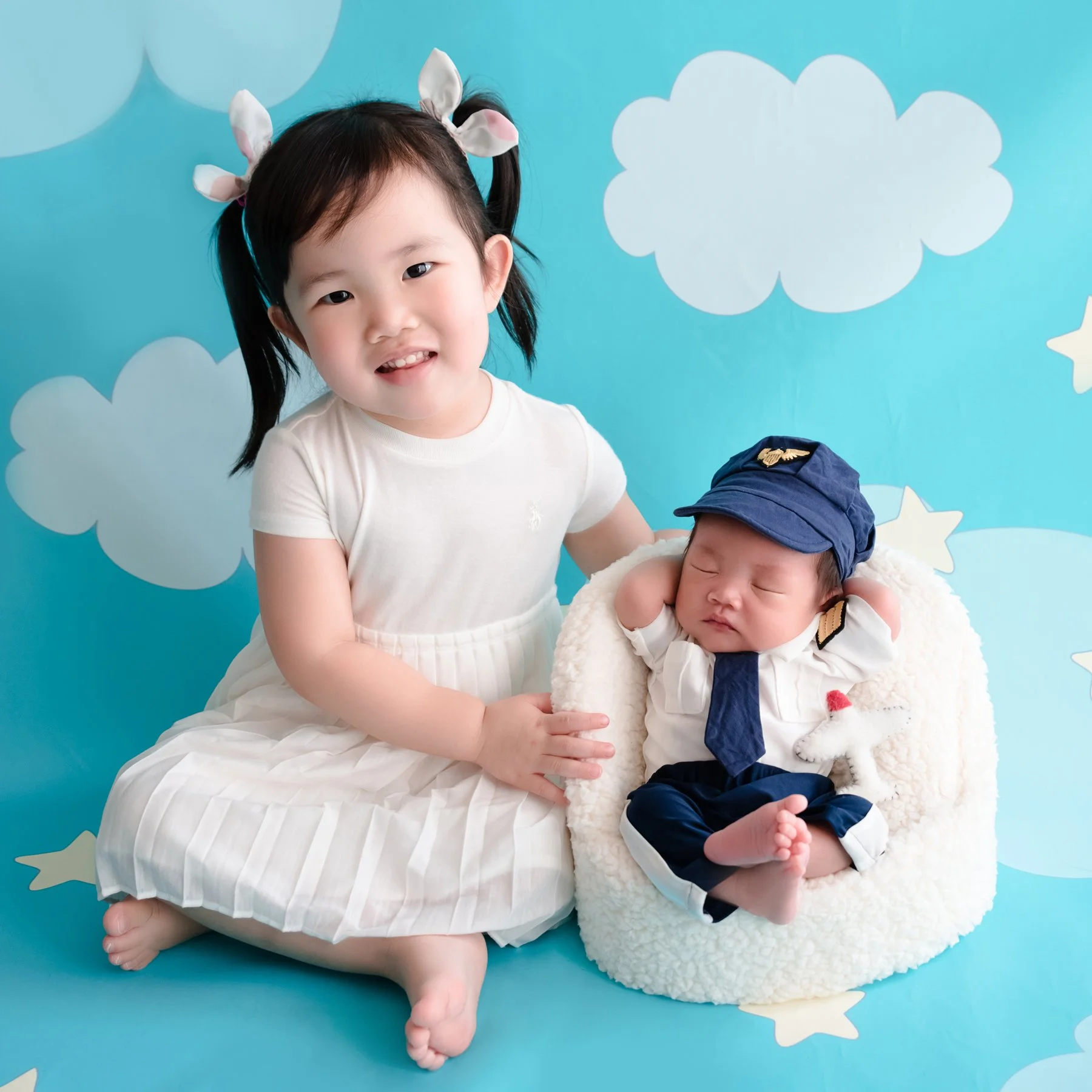 Older sister in white sitting beside her newborn sibling in a blue hat against a cloud backdrop