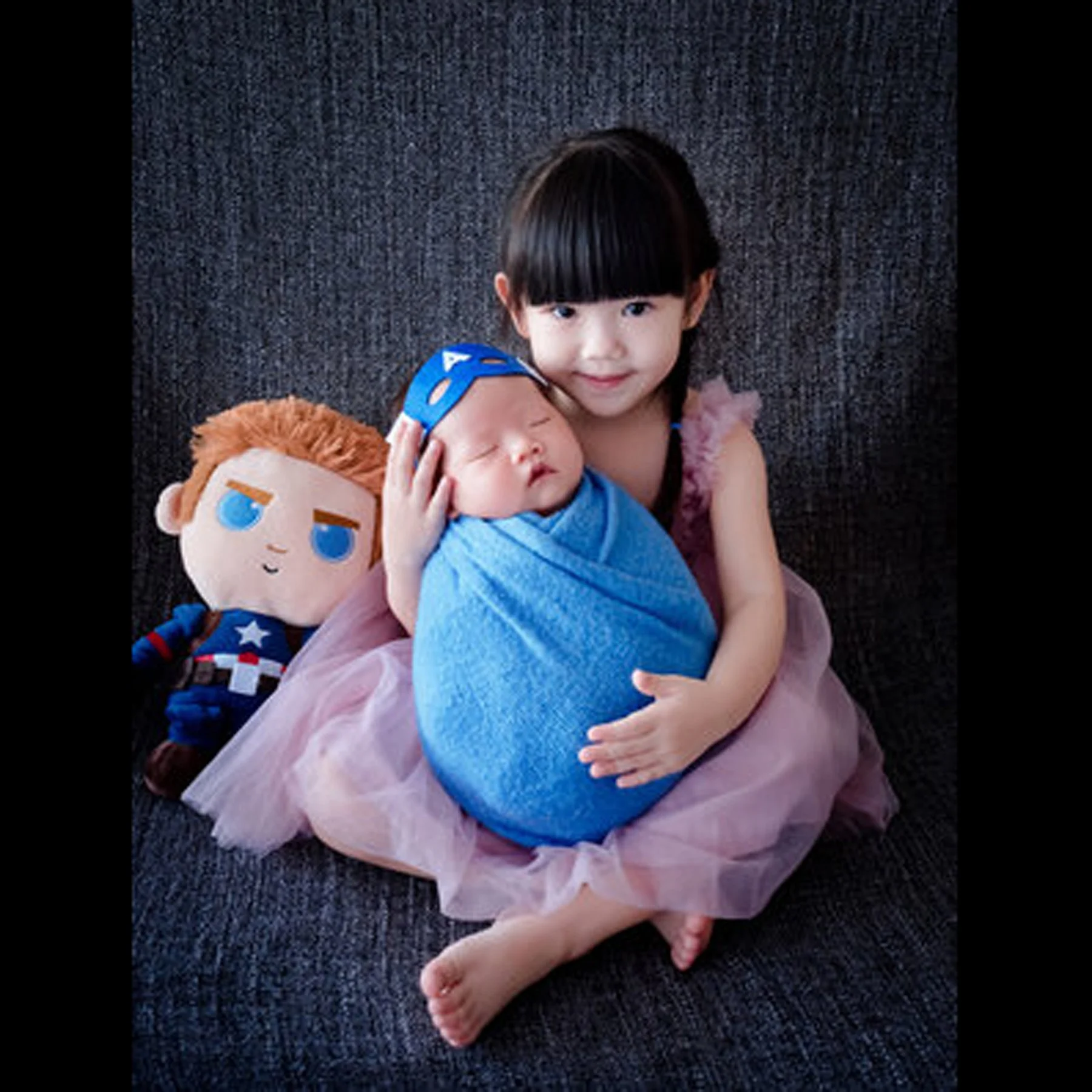 Older sister in a purple superhero outfit holding her newborn sibling wrapped in blue