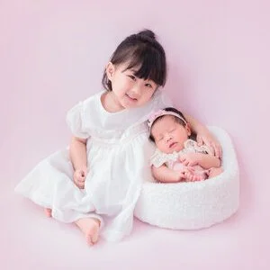 Older sister in a white dress sitting beside her newborn sibling in pink in a white basket