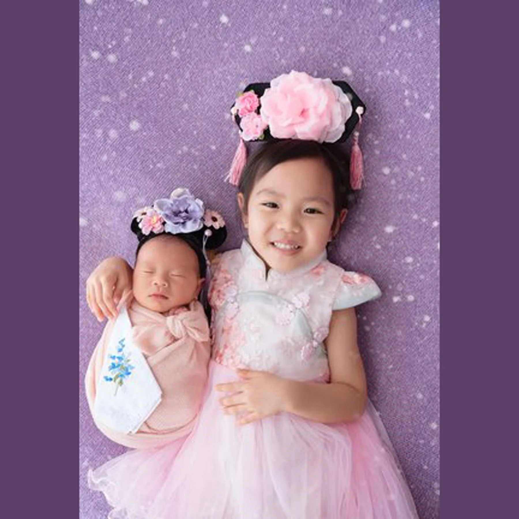 Older sister and newborn wearing floral headbands and pink outfits against a sparkling purple backdrop