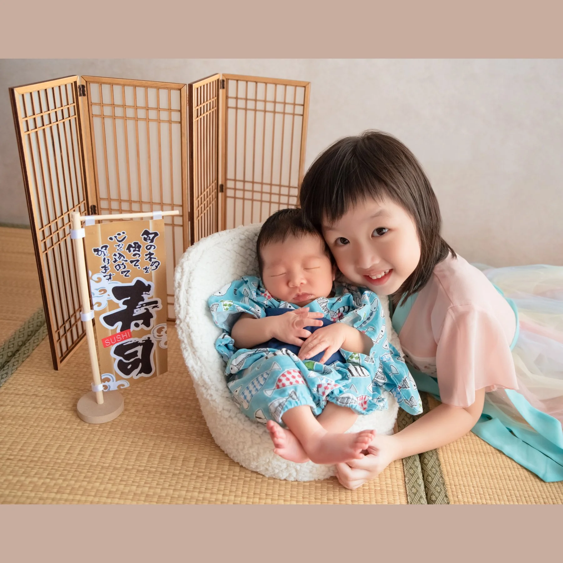 Older sister smiling beside her newborn sibling in a floral kimono and cream basket