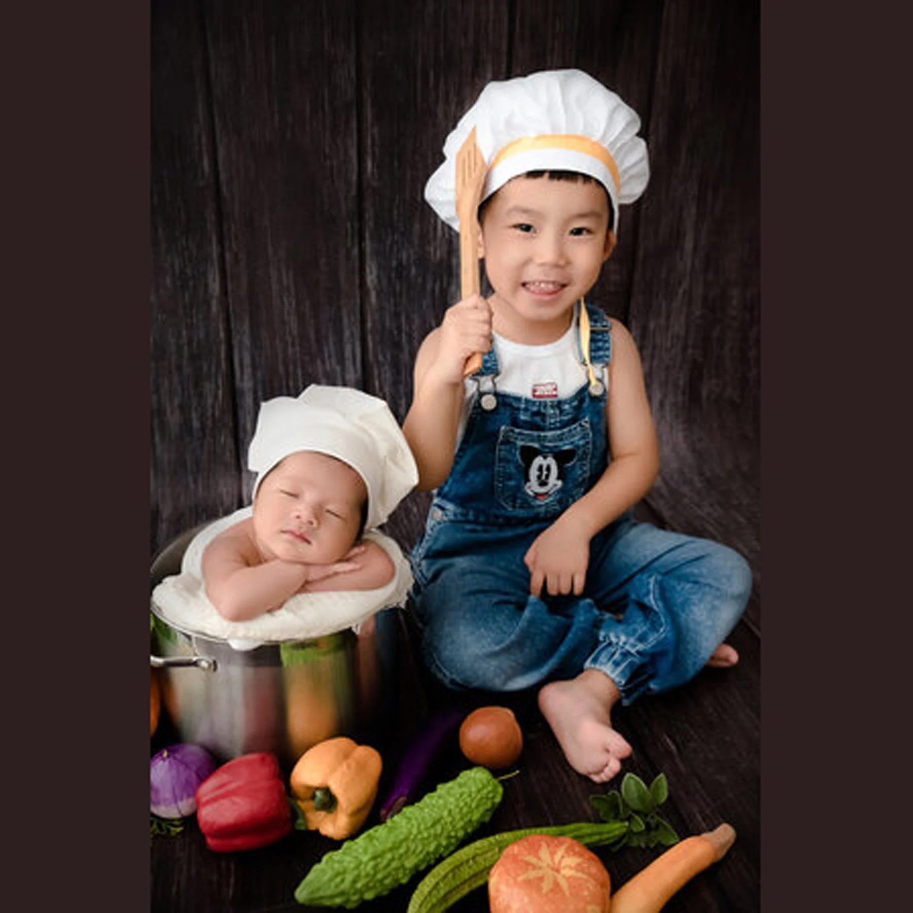 Older brother wearing a chef hat beside his sleeping newborn sibling and colourful vegetables
