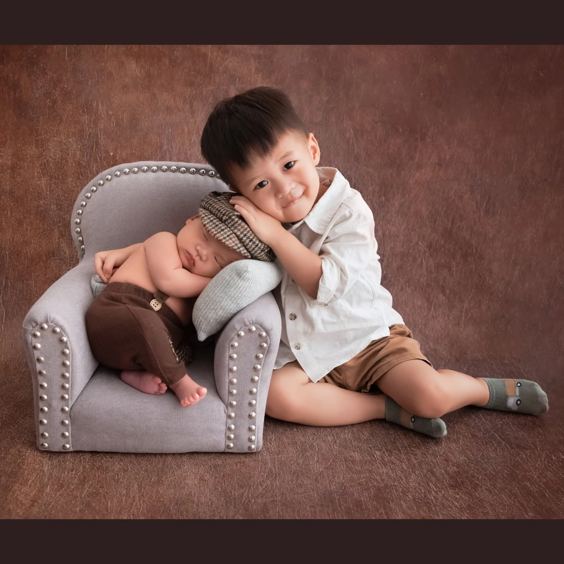 Older brother cuddling his newborn sibling while seated in a miniature grey armchair