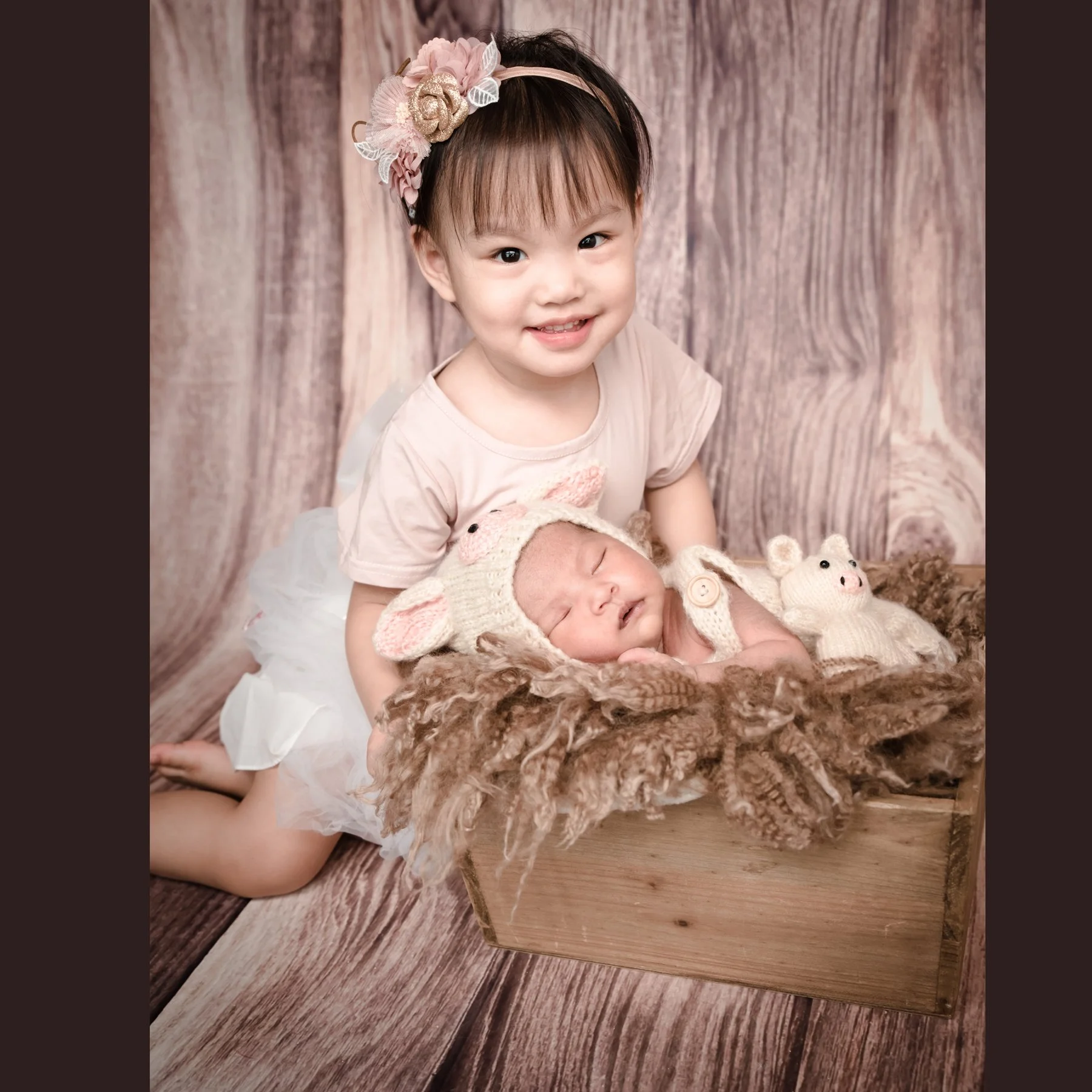 Older sister wearing a floral headband beside her newborn sibling in a rustic basket