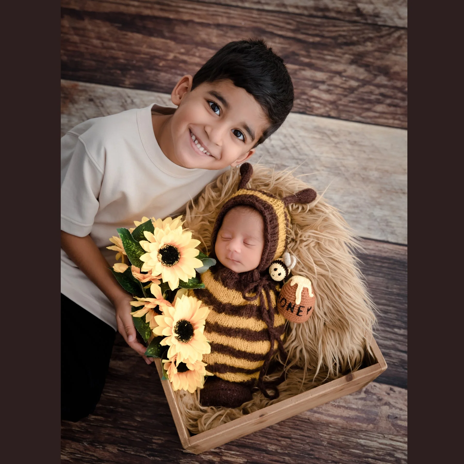 Older brother kissing his newborn sibling in a bee outfit beside sunflowers