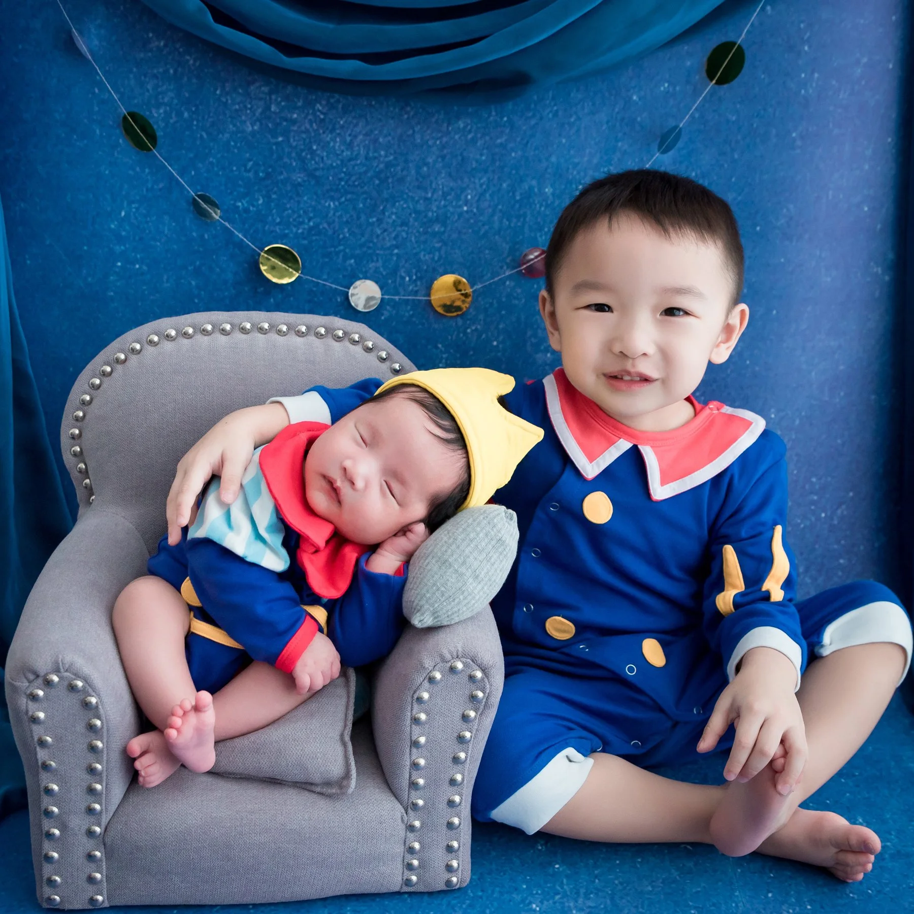 Older brother in a blue superhero outfit holding his newborn sibling dressed as Superman