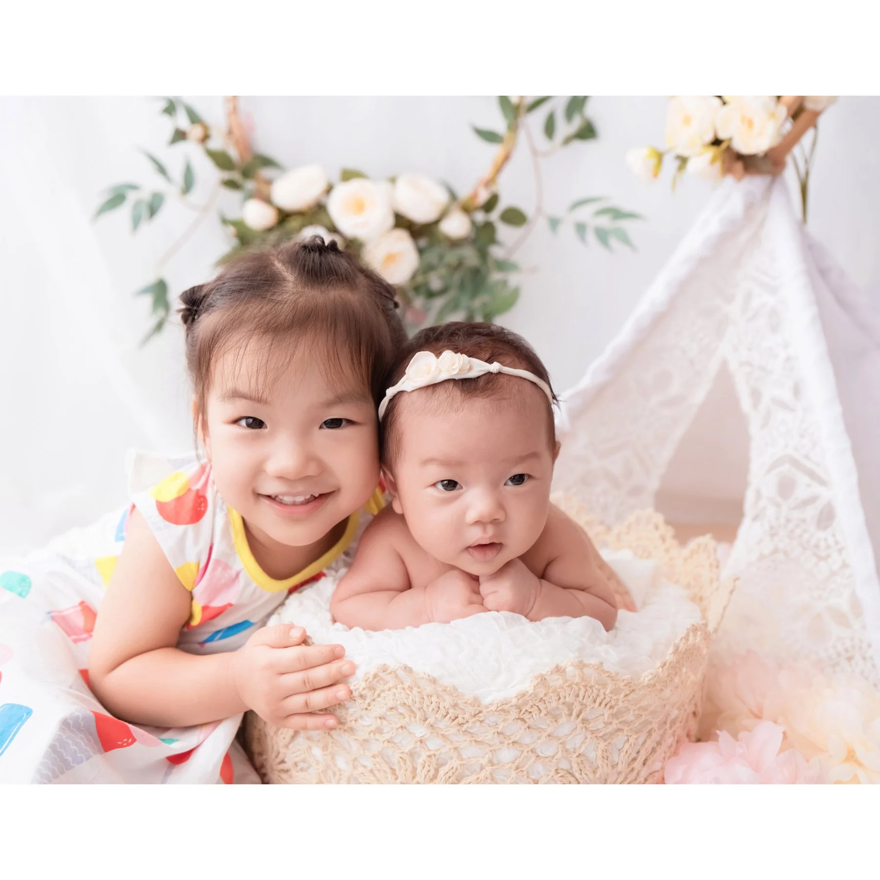 Older sister smiling beside her newborn sibling in a floral basket