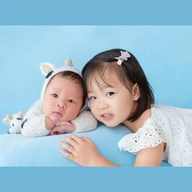 Older sister cuddling her newborn sibling in a white bear hat against a blue backdrop