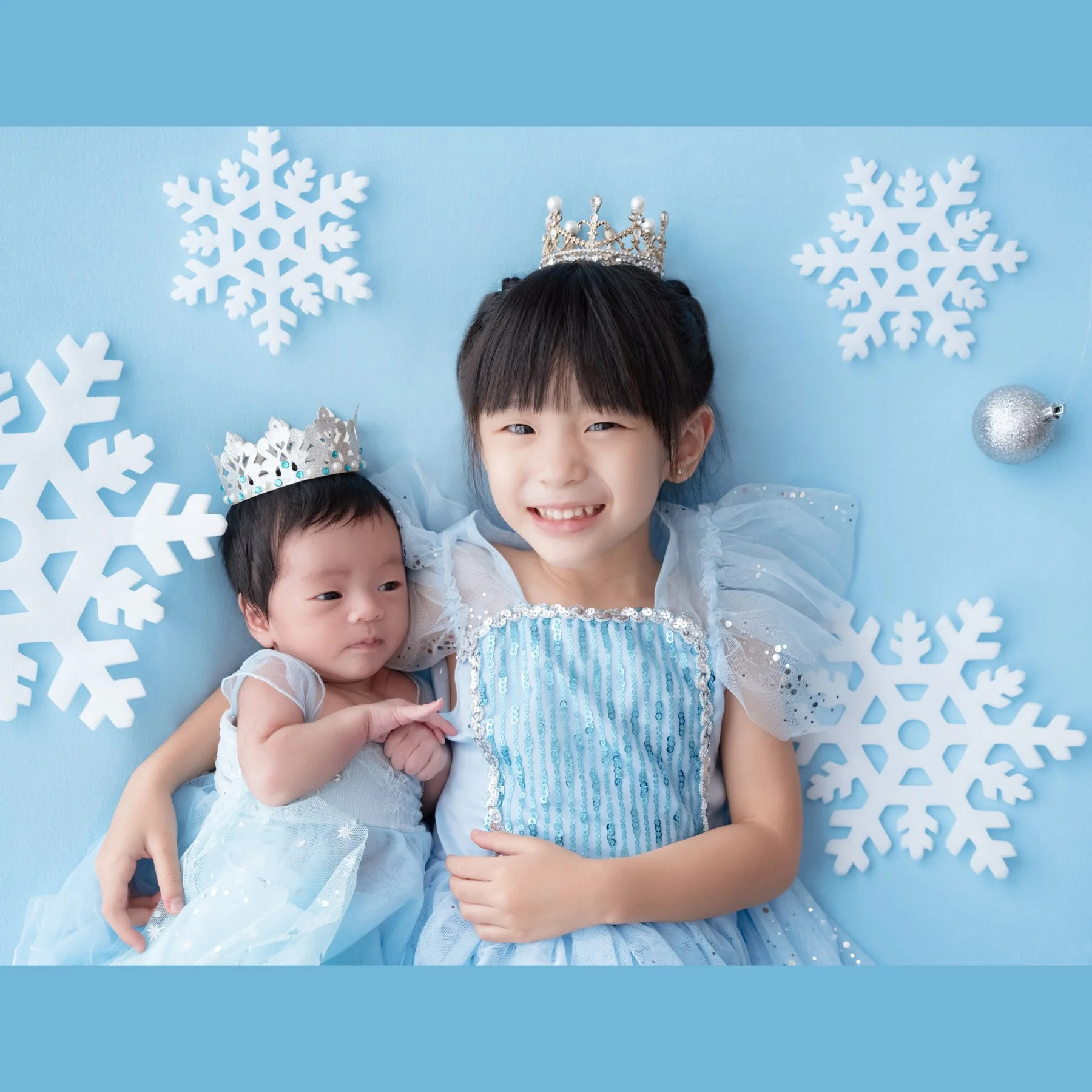 Older sister and newborn wearing blue outfits and tiaras against a snowflake backdrop