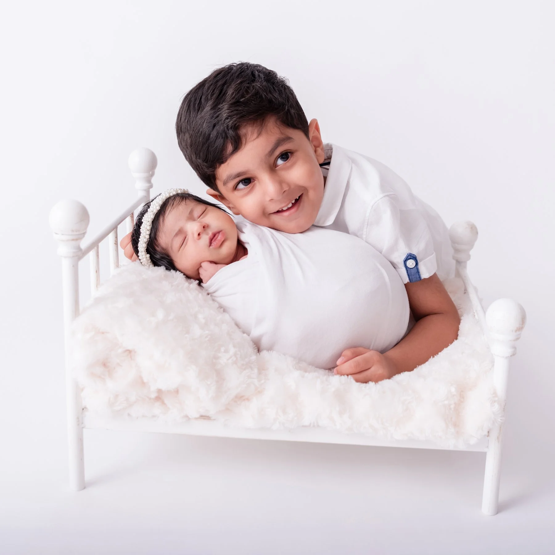 Older brother in a white shirt holding his swaddled newborn sibling on a white miniature bed