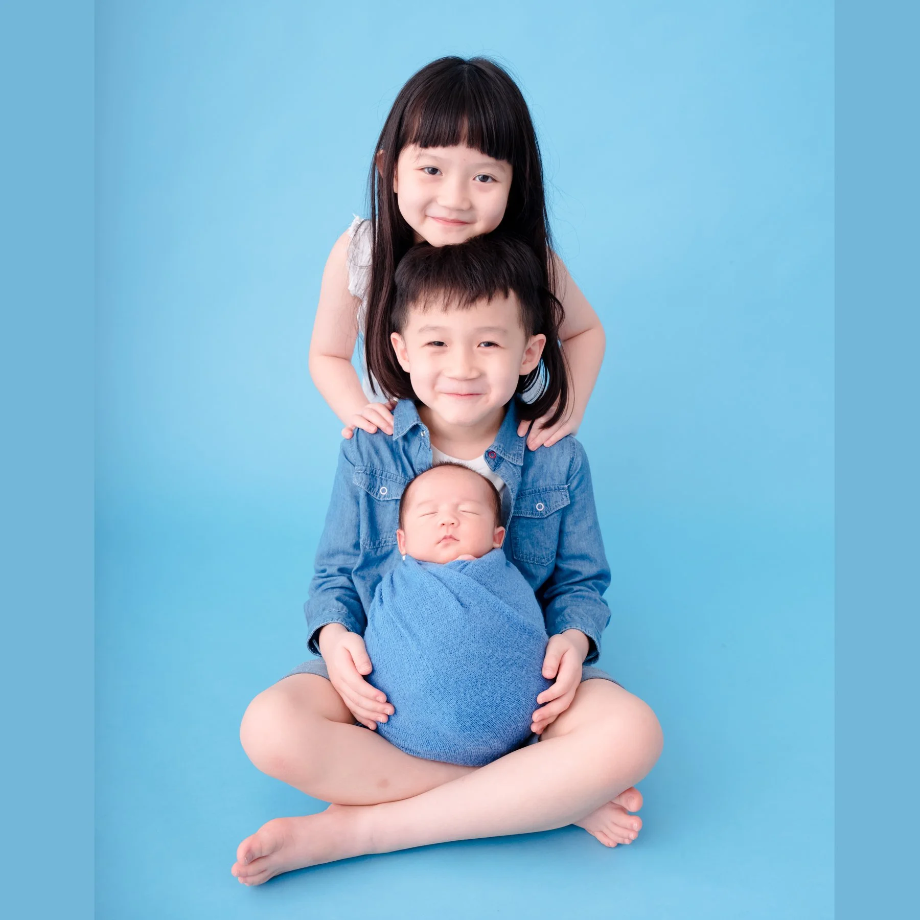 Older sister standing behind her brother as he holds their newborn sibling wrapped in blue