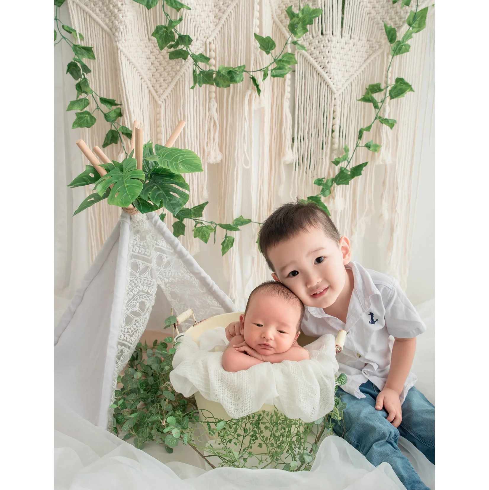 Older brother smiling beside his newborn sibling in a basket beneath a leafy woodland canopy