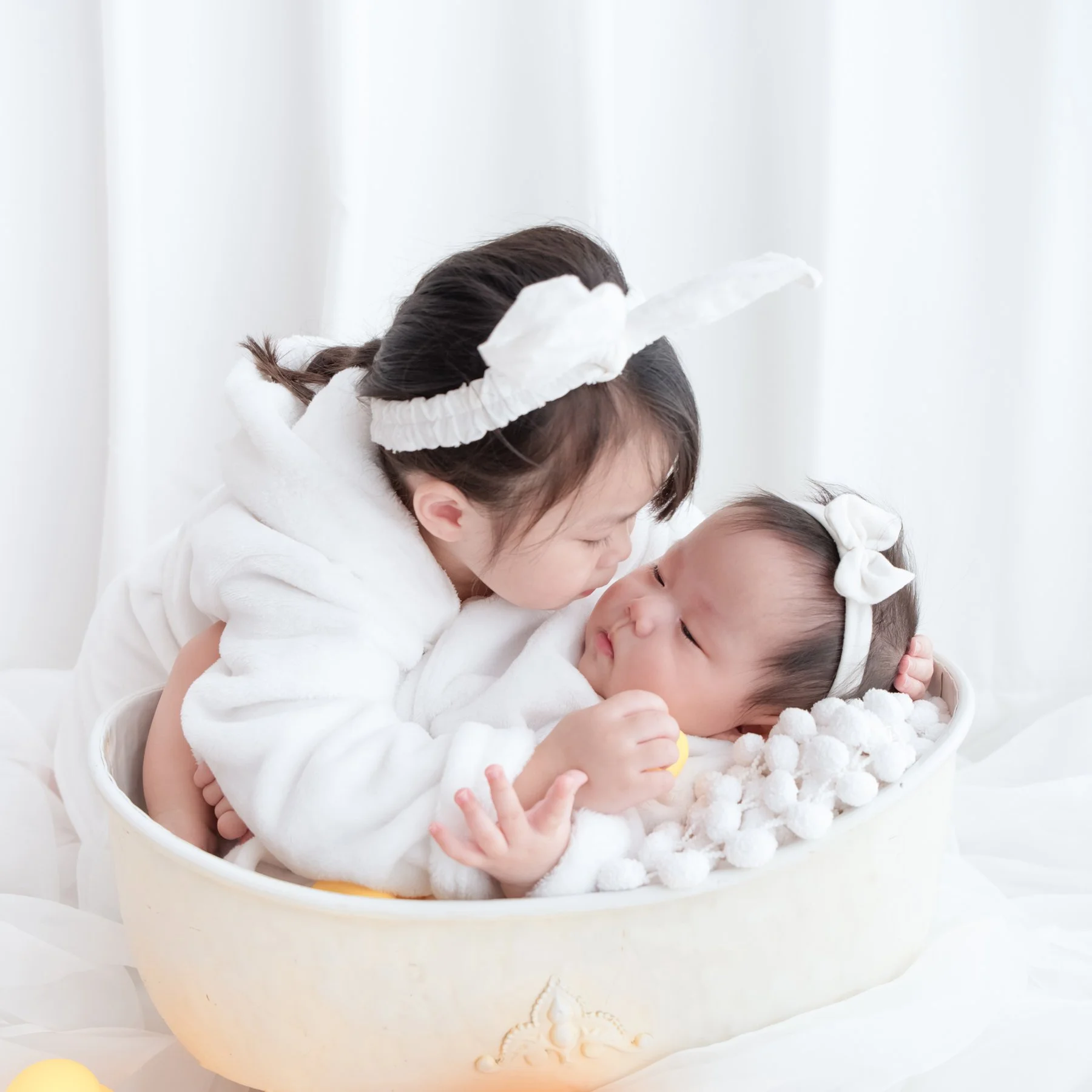 Older sister in a white bow cuddling her newborn sibling in a cream basket