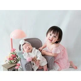Older sister in pink sitting beside her newborn sibling on a grey chair with flowers and pastel balloons