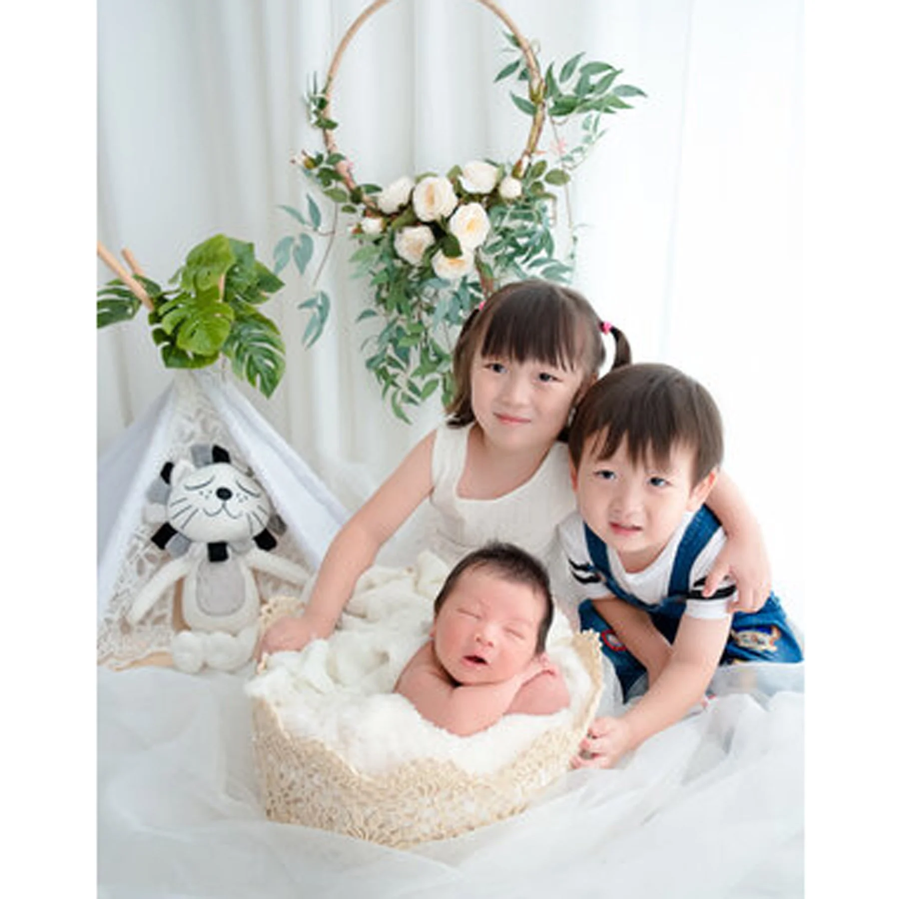 Older sister and brother leaning beside their newborn sibling beneath a leafy canopy