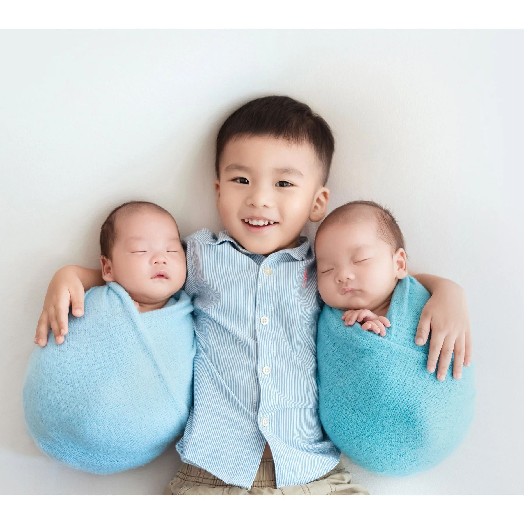 Older brother smiling between his newborn twin siblings wrapped in blue