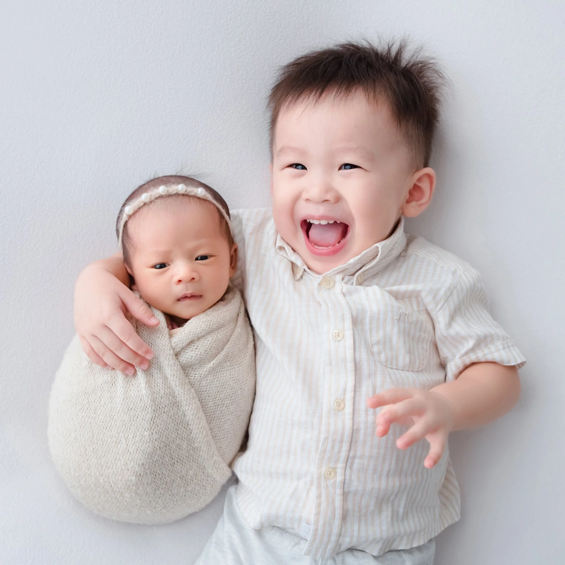 Smiling older brother sitting beside his swaddled newborn sibling in a small cream basket