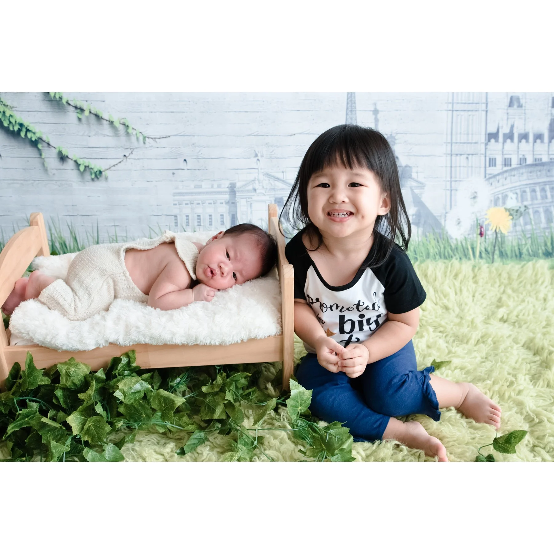 Older sister sitting beside her sleeping newborn sibling in a rustic basket on a woodland set