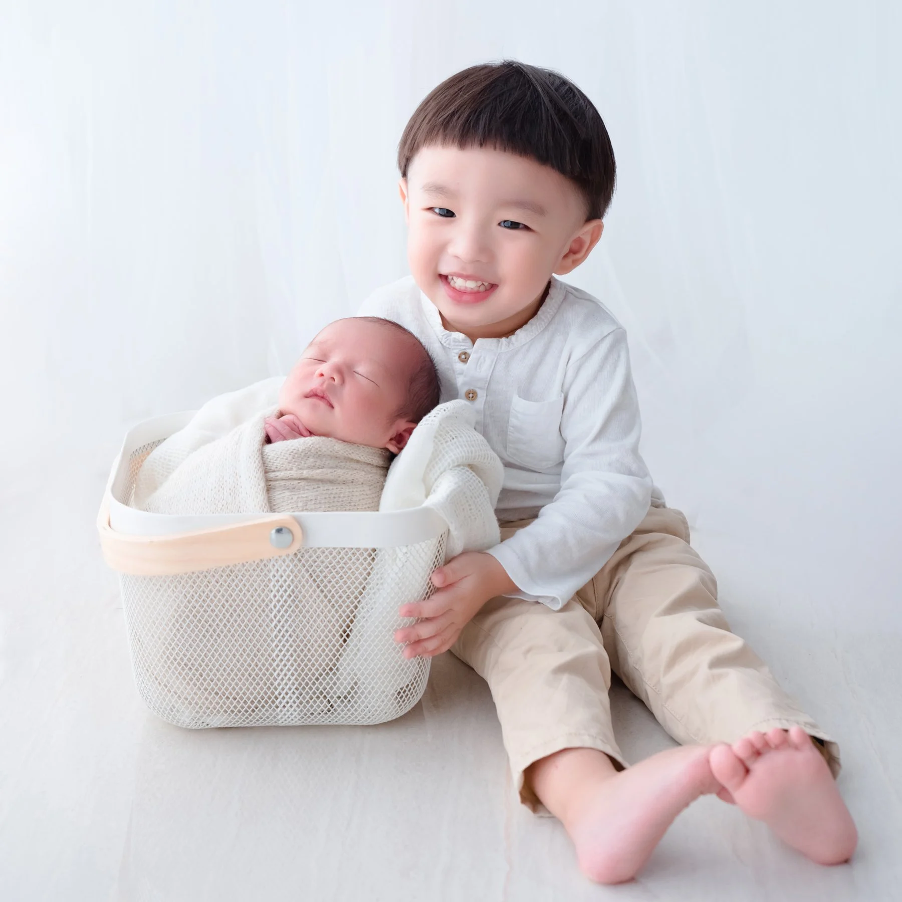 Older brother in white sitting beside his newborn sibling wrapped in cream in a white basket